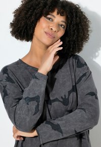 Donna con capelli ricci, leggeri efelidi, che indossa una felpa grigia con motivi di pipistrelli neri, appoggiando il viso su una mano, guardando leggermente verso l'alto.