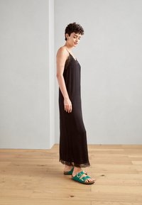 Svart slipklänning med spetskant och spagettiband, i kombination med turkosa sandaler med två spännen. Platt trägolv och neutral bakgrund.