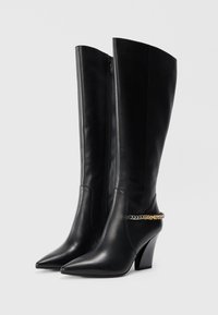 Knee-high black leather boots with pointed toes, a chunky heel, and a gold and silver chain accent around the ankle. Zipper closure.