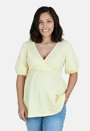 Smilende gravid kvinde med skulderlangt mørkt hår, iført en lysegul ammebluse og blå jeans, med hånd i lommen.