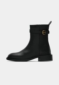 Černá kotníková bota s krokodýlím vzorem, nízkým robustním podpatkem, postranním páskem s přezkou a poutkem na zatahování na čistě bílém pozadí.