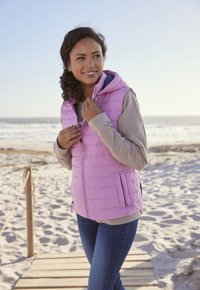 Hellrosa ärmellose Steppweste mit Kapuze, Reißverschluss vorne und seitlichen Taschen, getragen über einem grauen Langarmshirt; darunter sind Jeans sichtbar.