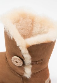 Botte en daim marron avec une doublure en shearling duveteuse couleur crème et un bouton décoratif sur le côté.