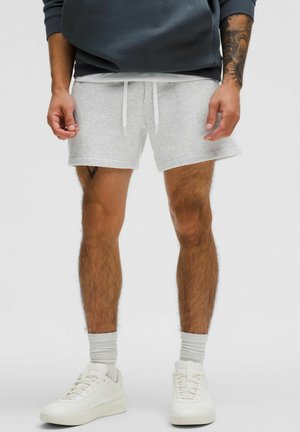 Man draagt lichtgrijze short met trekkoord, witte sokken en witte sneakers, staat met ontspannen handen langs het lichaam.