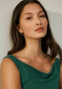 Mujer joven con cabello largo y oscuro, vestida con una blusa sin mangas verde y pendientes de aro dorados, mirando con confianza a la cámara frente a un fondo liso.