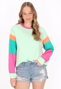 Maglietta verde chiaro a maniche lunghe con strisce rosa, arancioni e turchesi sulle maniche. Il materiale sembra morbido. Indossata con pantaloni shorts in denim strappati.