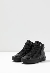 Svarte høye sneakers med en glinsende lakkfinish, kviltet tekstur på sidene, snøring foran og detalj med glidelås på siden.