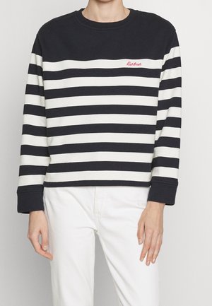 Un mannequin porte un haut à manches longues à rayures noires et blanches et un pantalon blanc sur un fond uni.