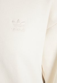 Bílá bavlněná mikina s malým, reliéfním logem Adidas na hrudi a měkkým, strukturovaným povrchem.