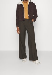 Persona che indossa una giacca di velluto a coste marrone con fodera bianca, una camicia senape, pantaloni marrone scuro larghi e sneakers bianche, in piedi con una mano in tasca.