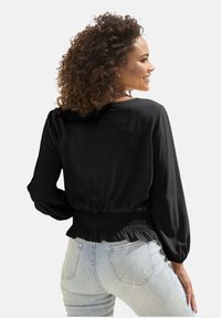 Schwarze Bluse mit langen Ärmeln und gesmoktem Bund, mit glatter Textur. Kombiniert mit hellblauen, verwaschenen Jeans. Rückansicht gezeigt.