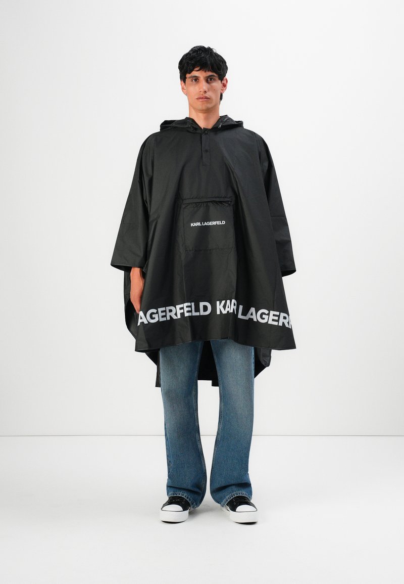 Man in een zwarte Karl Lagerfeld regenponcho met capuchon, over blauwe jeans en zwart-witte sneakers, staande voor een witte achtergrond.