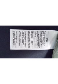 Etichetta con istruzioni per la cura e composizione del materiale: 84% cotone, 16% poliestere. Testo multilingue con intestazioni chiare e dettagli sul tessuto.