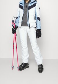 Lyžařský outfit s modro-bílou péřovou bundou, šedým trikem, bílými kalhotami, černými rukavicemi a černými lyžařskými botami, držící růžové lyžařské hůlky.