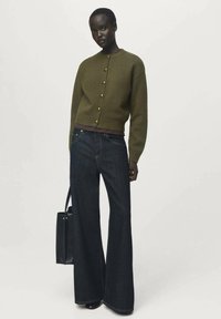 Cardigan de punto verde oliva con botones en la parte delantera, combinado con pantalones vaqueros de pierna ancha en azul oscuro. La modelo sostiene un bolso tote negro.