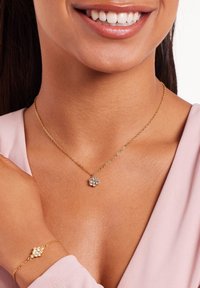 Gouden ketting met een bloemenhanger met een cluster van heldere stenen. Armband met een vergelijkbaar ontwerp, gedragen om de pols. Geplaatst tegen een lichtroze top.