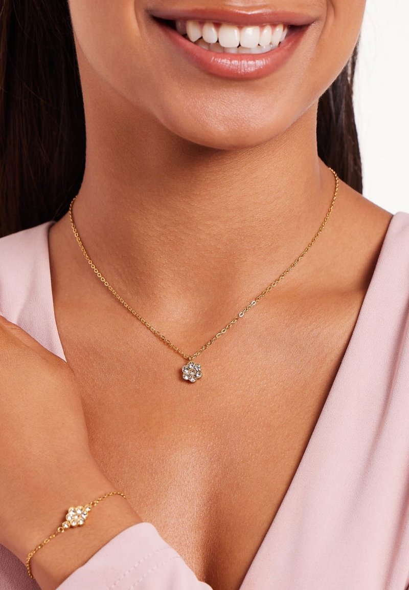 Gouden ketting met een bloemenhanger met een cluster van heldere stenen. Armband met een vergelijkbaar ontwerp, gedragen om de pols. Geplaatst tegen een lichtroze top.