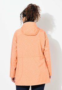 Chaqueta impermeable ligera en melocotón con lunares blancos, que cuenta con capucha, bolsillos laterales y un diseño de corte recto.