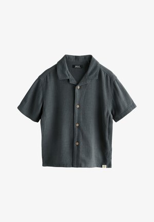 Jeune enfant aux cheveux foncés portant une chemise sombre à manches courtes boutonnée et un short clair, regardant vers le bas les bras légèrement tendus.
