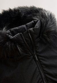 Veste noire avec un col en fausse fourrure, une fermeture éclair à l'avant et un tissu texturé qui semble léger et résistant à l'eau.