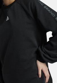 Sudadera negra con cuello redondo y mangas largas. Presenta el logo de Adidas en blanco y un detalle de texto en negrita en la manga. Tejido suave.