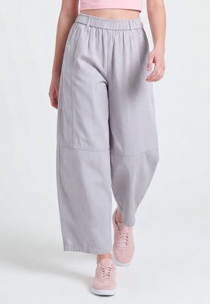 Person trägt lockere, hellgraue, weit geschnittene Hosen mit elastischem Bund und pinken Sneakers und geht vor weißem Hintergrund.