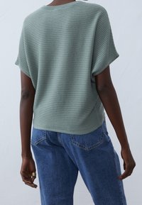 Osoba ubrana w zielony top z krótkim rękawem w prążki i niebieskie dżinsy, widoczna od tyłu, z jedną ręką swobodnie opuszczoną i złotym pierścionkiem na palcu.