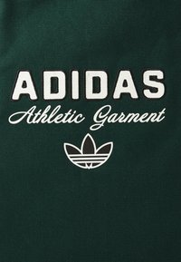 Dunkelgrüner Stoff mit weißem "ADIDAS Athletic Garment"-Text und einem schwarz-weißen Adidas-Trefoil-Logo unter dem Text.