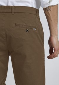 Pantalons en coton marron avec poche arrière dotée d'une fermeture à bouton et d'un accent rayé. La vue latérale montre une texture lisse et une coupe ajustée.