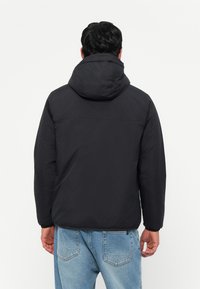 Veste rembourrée noire avec capuche, offrant une texture lisse et des poignets élastiques. La vue arrière montre un design simple sans logos ni motifs visibles.
