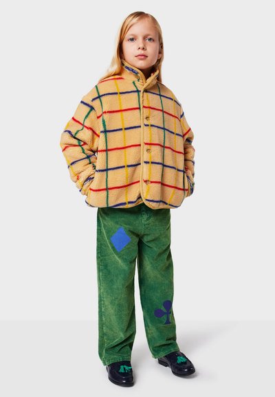 Giacca di pile gialla a scacchi con linee rosse, blu e verdi. Pantaloni in corduroy verde con toppe a diamante blu e trifoglio, scarpe nere.