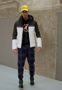 Chaqueta acolchada en blanco y negro, sudadera negra con logo naranja, pantalones a cuadros en azul y verde, y zapatillas verdes con suelas blancas. Gorro amarillo.