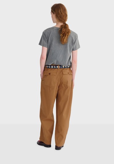 Personne aux cheveux roux attachés en queue de cheval, portant un t-shirt gris délavé à manches courtes, un pantalon marron large avec des poches arrière à boutons, et des chaussures foncées.