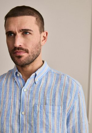 Man with short brown hair and beard wearing a light blue, buttoned striped shirt with a chest pocket, looking slightly left on plain background.
