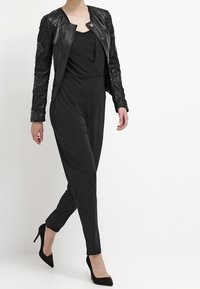 Chaqueta de cuero negra con mangas texturizadas, combinada con un mono negro. Los tacones altos completan el elegante atuendo monocromático.