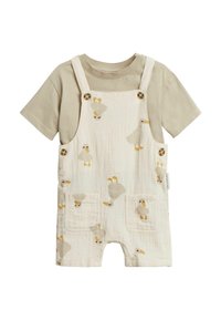 Korte overall romper in crème met eendenpatronen, voorzien van houten knopen en twee voorzakken. Vergezeld van een beige t-shirt eronder.