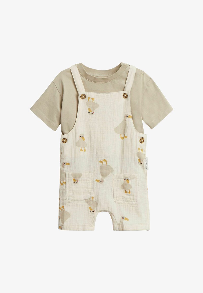 Kurzer Overall in Creme mit Entenmustern, ausgestattet mit Holzknöpfen und zwei vorderen Taschen. Dazu wird ein beiges T-Shirt darunter getragen.