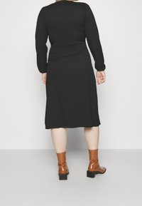Vestido negro de manga larga con corsé ajustado y falda acampanada, combinado con botas de tobillo brillantes marrones con tacón cuadrado.