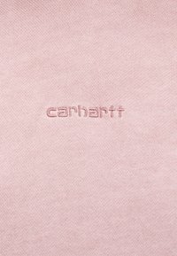 Világos rózsaszín pamutanyag, amelyen domború "carhartt" logó található. A textúra sima és egységes, további minták vagy kiemelések nélkül.