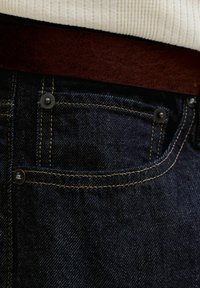 Κοντινό πλάνο στην τσέπη σκούρου μπλε τζιν με κίτρινη ραφή, καφέ δερμάτινη ζώνη και λευκή ριγωτή άκρη πουκαμίσου.