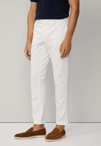 Pantaloni bianchi slim-fit in cotone con una texture liscia, dotati di tasche laterali e caviglie affusolate. Abbinati a scarpe slip-on marroni.