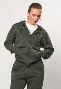 Frau mit zurückgebundenen Haaren, die goldene Creolen trägt, olivgrüner Kapuzenpullover mit Reißverschluss, passende Hose und weißes T-Shirt mit Aufdruck, Hände in den Taschen.