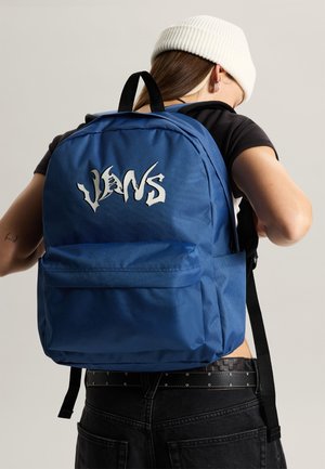 Person iført hvid hue og sort t-shirt, der justerer en blå Vans rygsæk med forlomme og sorte stropper mod en ensfarvet baggrund.