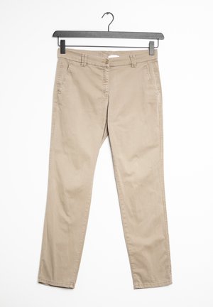Beige chino-bukser med sidelommer og bæltestropper hængende på en sort bøjle mod en hvid baggrund.