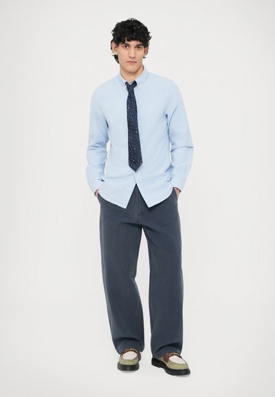 Vaaleansininen nappipaita, jossa on kaulus, yhdistetty tummanharmaisiin löysästi istuviin housuihin ja ruskeilla yksityiskohdilla varustettuihin kenkiin. Mukana pilkullinen tumma solmio.