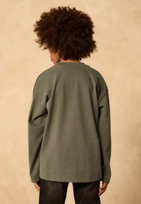 Bluză cu mâneci lungi, de bumbac verde măsliniu, cu croială lejeră, decolteu rotund și cusături căzute la umeri. Textură moale, design minimalist.