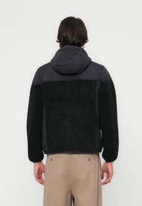 Veste en polaire noire doublée, avec capuche, présentant un panneau supérieur lisse noir en contraste. Matériau texturé au dos et sur les manches.