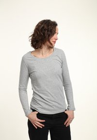 Helles graues Langarmshirt mit rundem Ausschnitt, aus weichem Stoff. Das Modell posiert mit den Händen in die Hüften gestützt, während es schwarze Hosen trägt.
