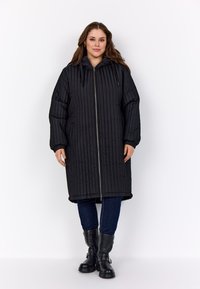 Nő áll egy hosszú fekete steppelt kabátban, kapucnival, sötétkék farmerban és fekete térd középig érő csizmában, fehér háttér előtt.