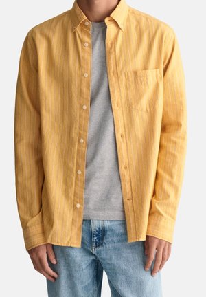 Hombre con una camisa amarilla de rayas desabrochada de manga larga sobre una camiseta gris y vaqueros azul claro, de pie frente a un fondo liso.